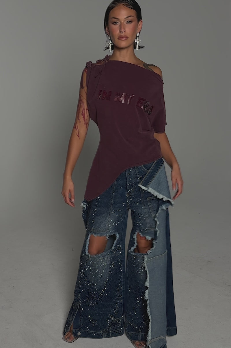 A video of a person in the IN MY ERA OFF-THE-SHOULDER TEE and wide-leg, ripped, embellished blue jeans, accessorized with tattoos, earrings, and slicked-back hair against a gray backdrop.