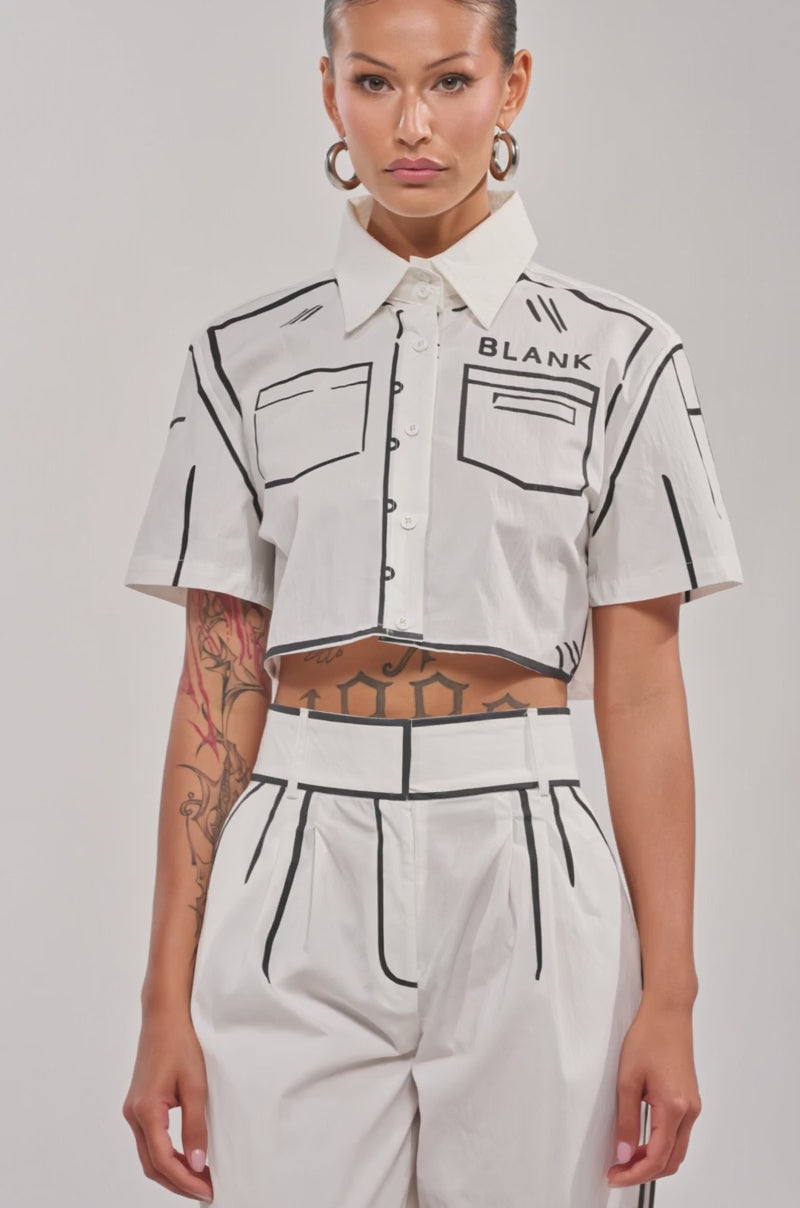 Video of woman modeling the BLANK SPACE Cropped Short Sleeve Button Down, a cotton blend pop-art shirt with bold black line designs and “BLANK” on the chest, plus matching high-waisted pants, hoop earrings, and arm tattoos.