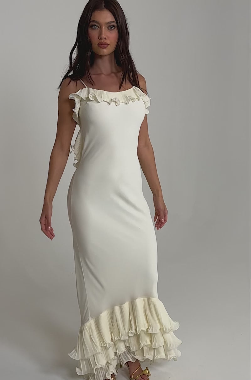 A video of a woman wearing the LOVE ME OR LOSE ME RUFFLE MAXI DRESS IN IVORY, a sleeveless maxi with layered ruffles at the hem, paired with gold sandals.