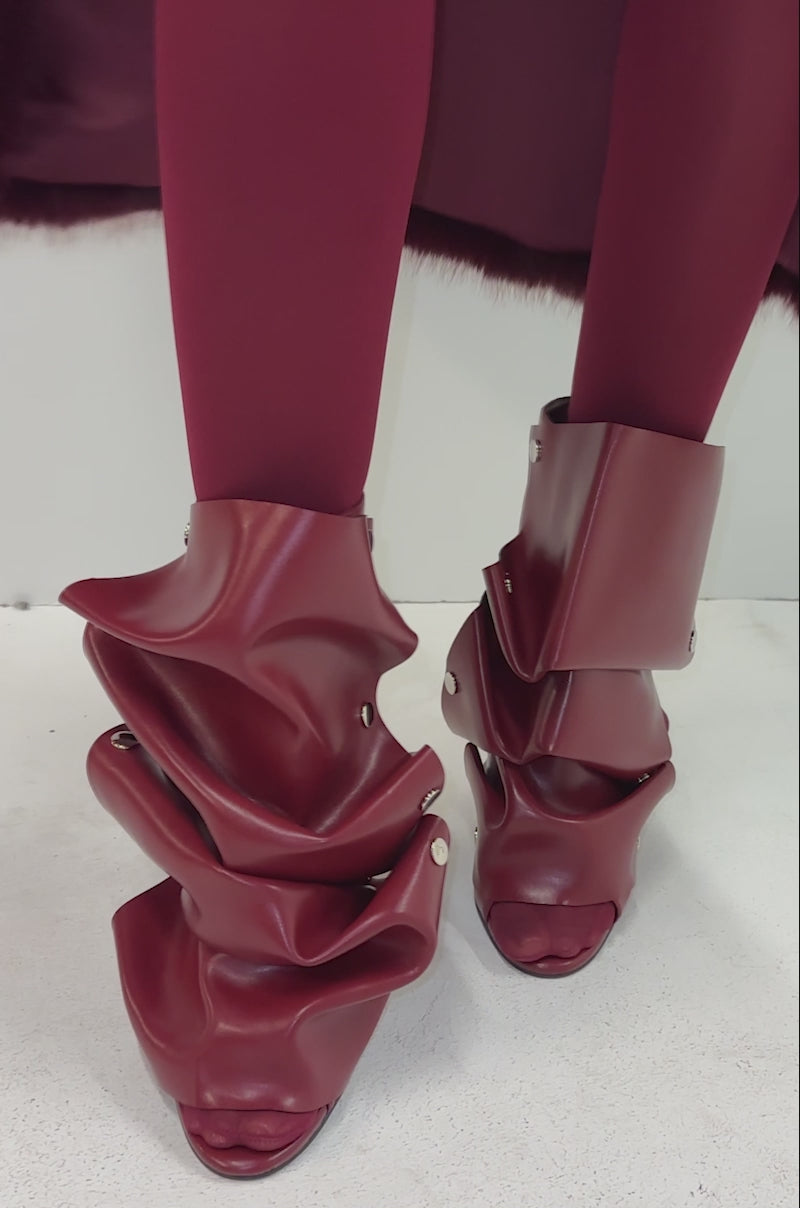 A video of a model in burgundy tights and a matching coat stands against a white background, showcasing the AZALEA WANG BIRDSONG Burgundy Snap Open Toe Stiletto Bootie with ruched, shiny leather and metallic studs.