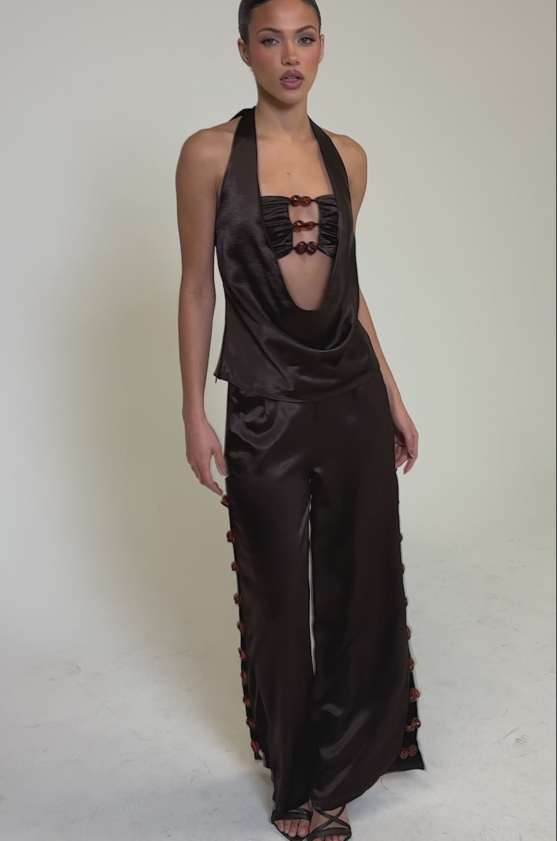 Video of woman modeling the RESORT VIBES TROUSER, a dark brown satin wide-leg pant, paired with a matching shiny halter top featuring a draped neckline and red accents. She completes the look with open-toe heels against a plain light background.
