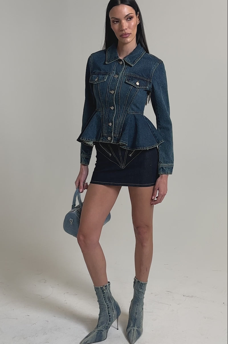 A video of a woman wearing the POP OUT PEPLUM DENIM JACKET with dark denim shorts, pointed denim heeled boots, and holding a small light blue denim handbag.