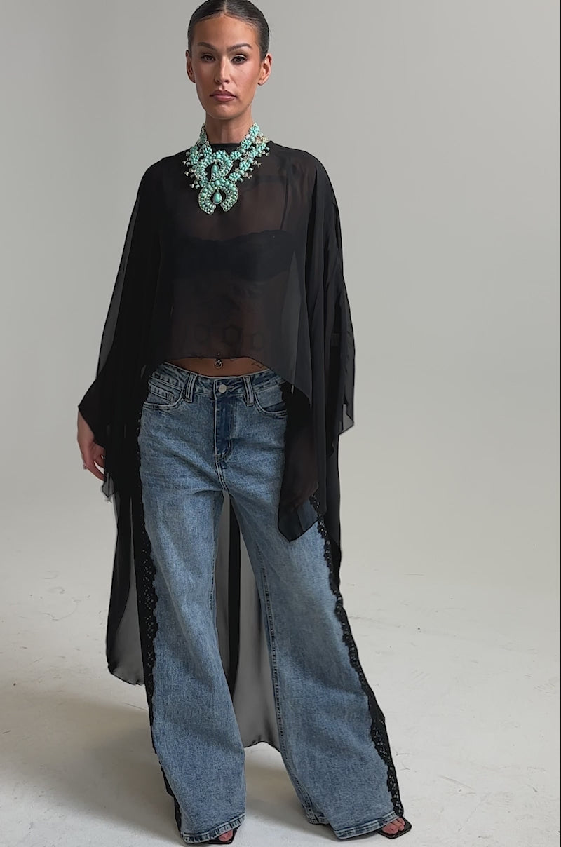 Video of woman modeling the COCO BLOUSE—a sheer chiffon top with cape sleeves—styled over a black bralette, wide-leg blue jeans with black lace sides, open-toe heels, and a bold turquoise necklace against a plain background.