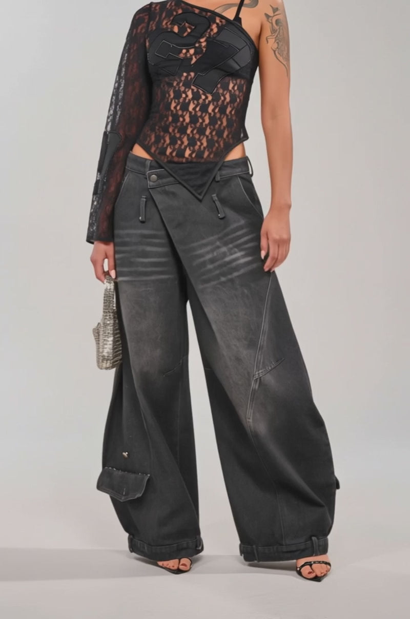 A video of a woman in the IZZY RELAXED DENIM IN BLACK WASH wide-leg pants, styled with a sheer black asymmetrical lace one-sleeve top, and a small silver handbag.