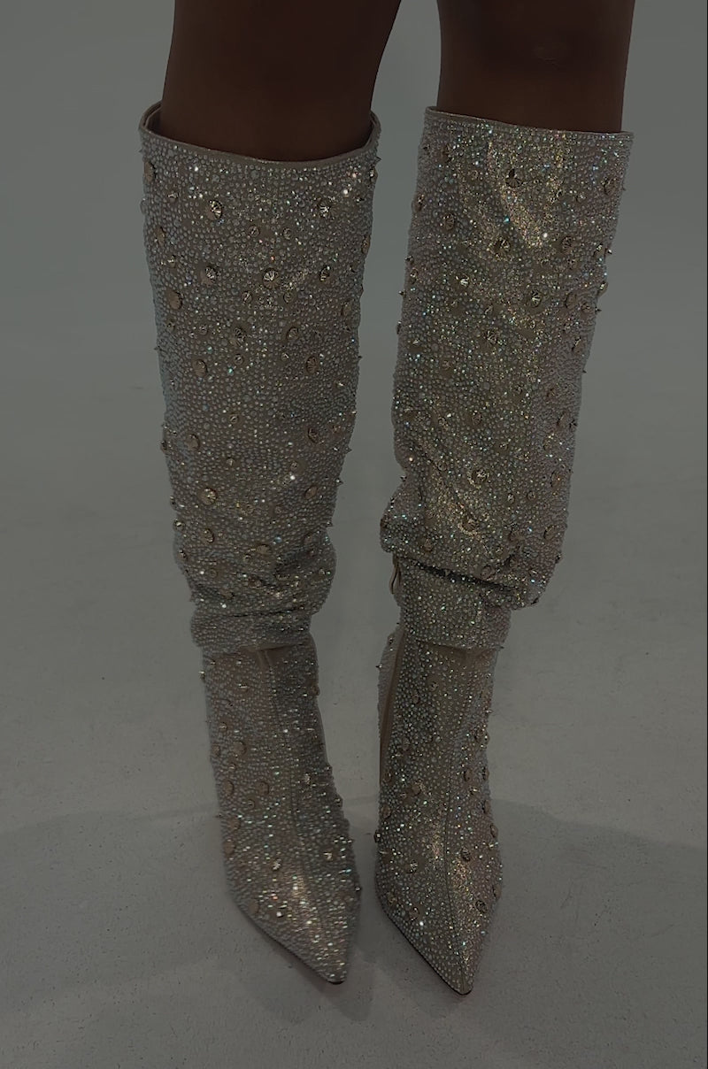 A video of a model wearing a shiny metallic mini skirt, a person stands on a light floor against a neutral background, showcasing the glamorous AZALEA WANG DOUGHTY GOLD STILETTO BOOTS.