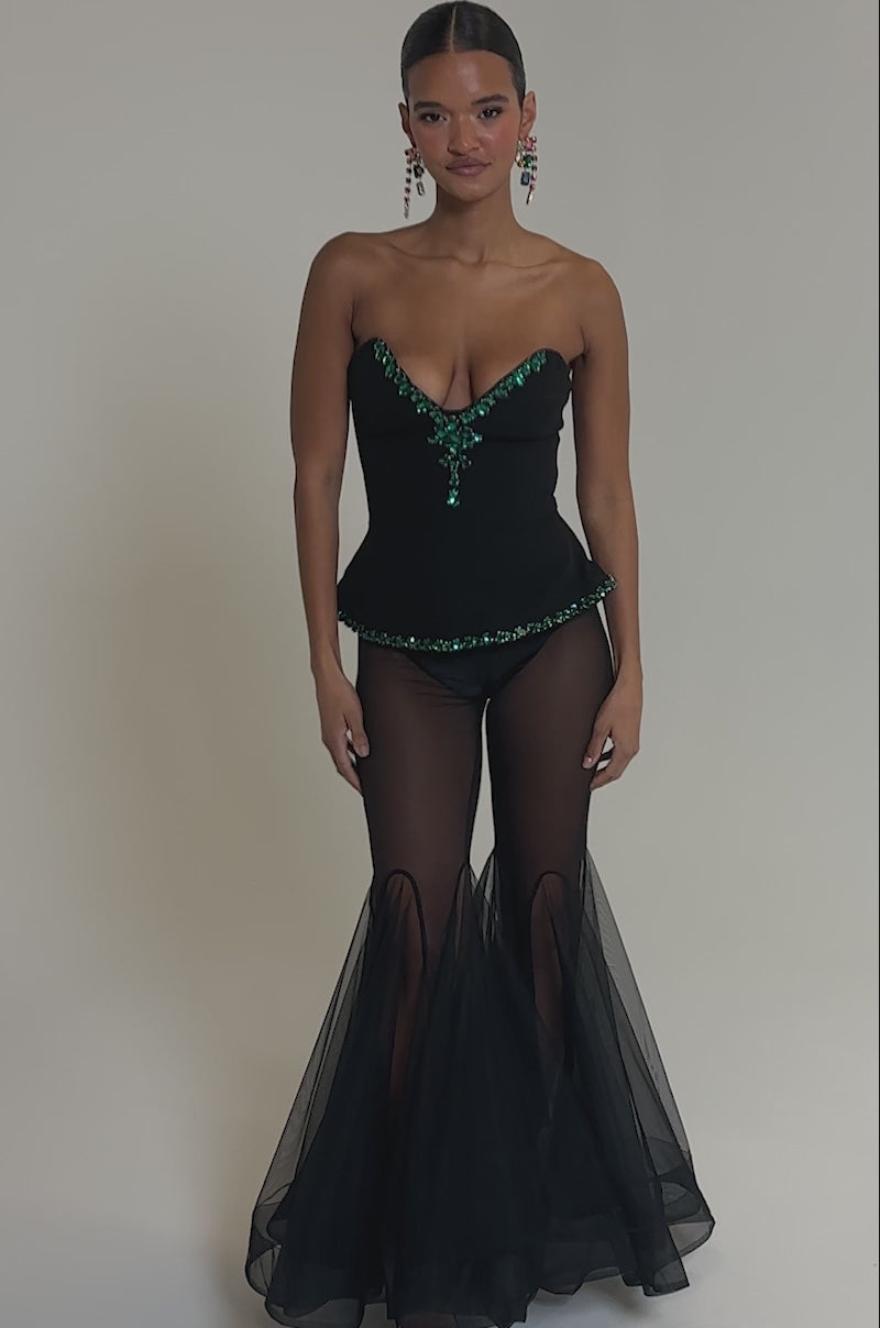 Video of woman modeling the JEWELS FOR EVERYONE CORSET, a strapless bejeweled top with emerald accents, matching earrings, and sheer black peplum pants, set against a plain gray background.