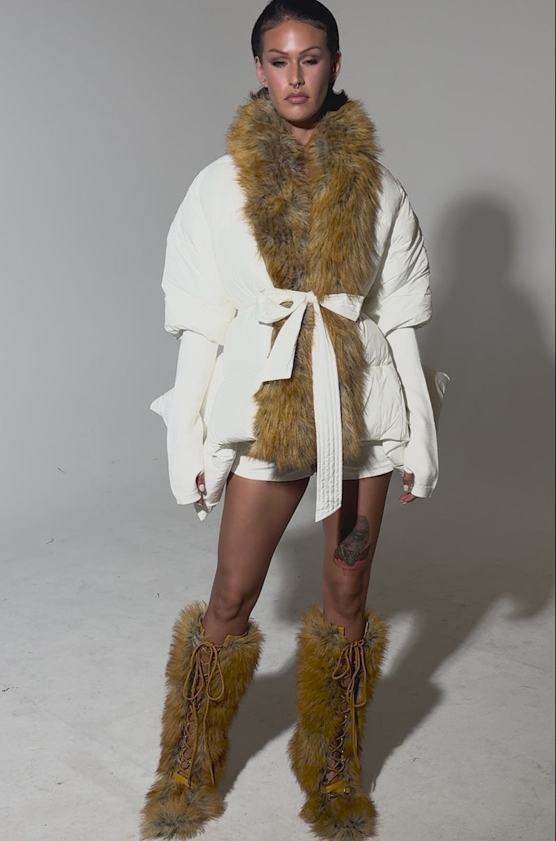 A video of a woman modeling the TALK TO ME NICE FUR TRIM PUFFER with a faux fur collar, paired with matching faux fur boots, white shorts, and shows off a thigh tattoo against a plain light backdrop.