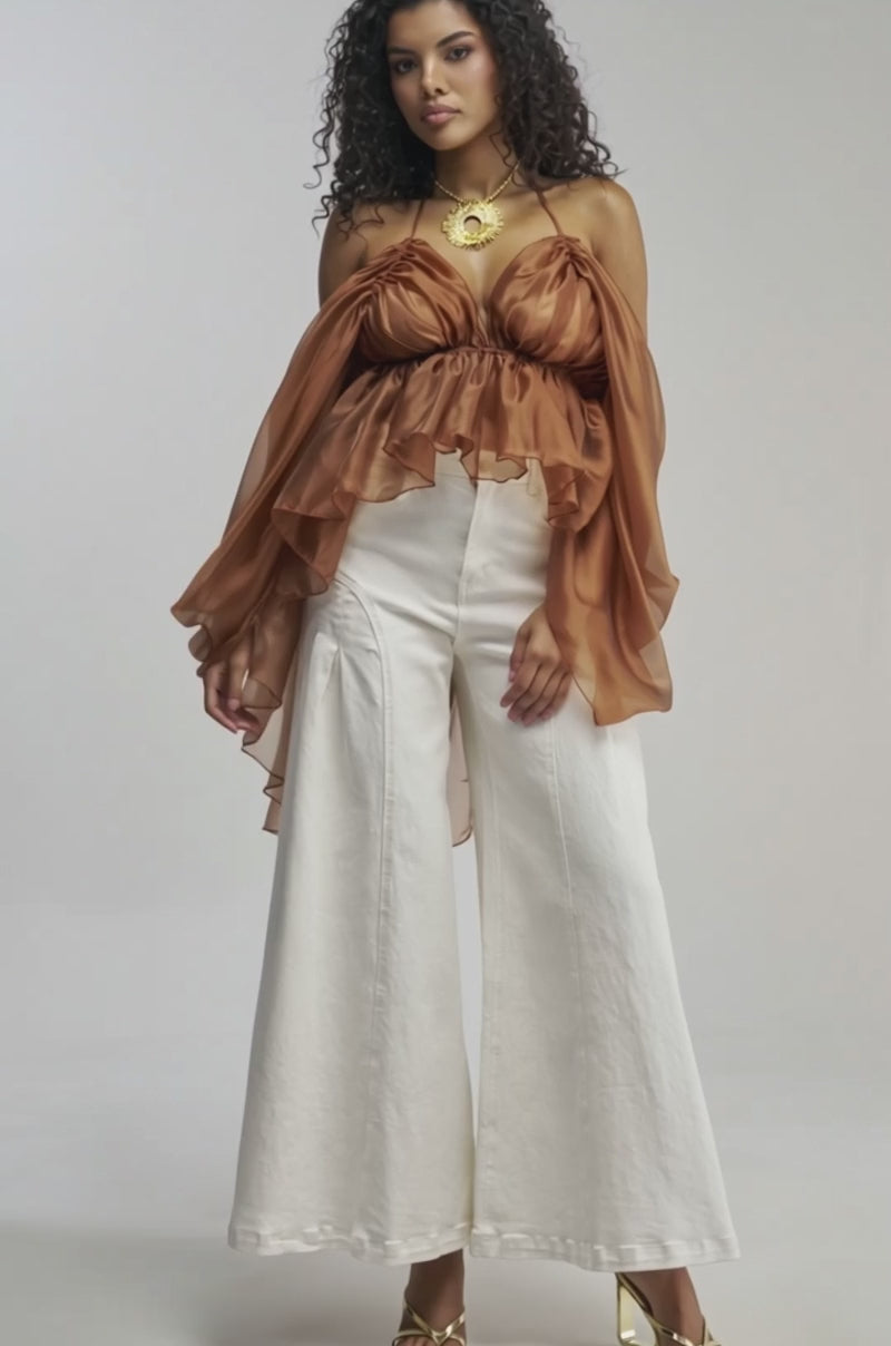 Video of woman modeling the FLY WITH ME CHIFFON BLOUSE—a rust-colored, open-back design featuring dramatic ruffled sleeves—paired with a statement necklace and high-waisted white pants, making it the perfect choice for standout occasion dressing.