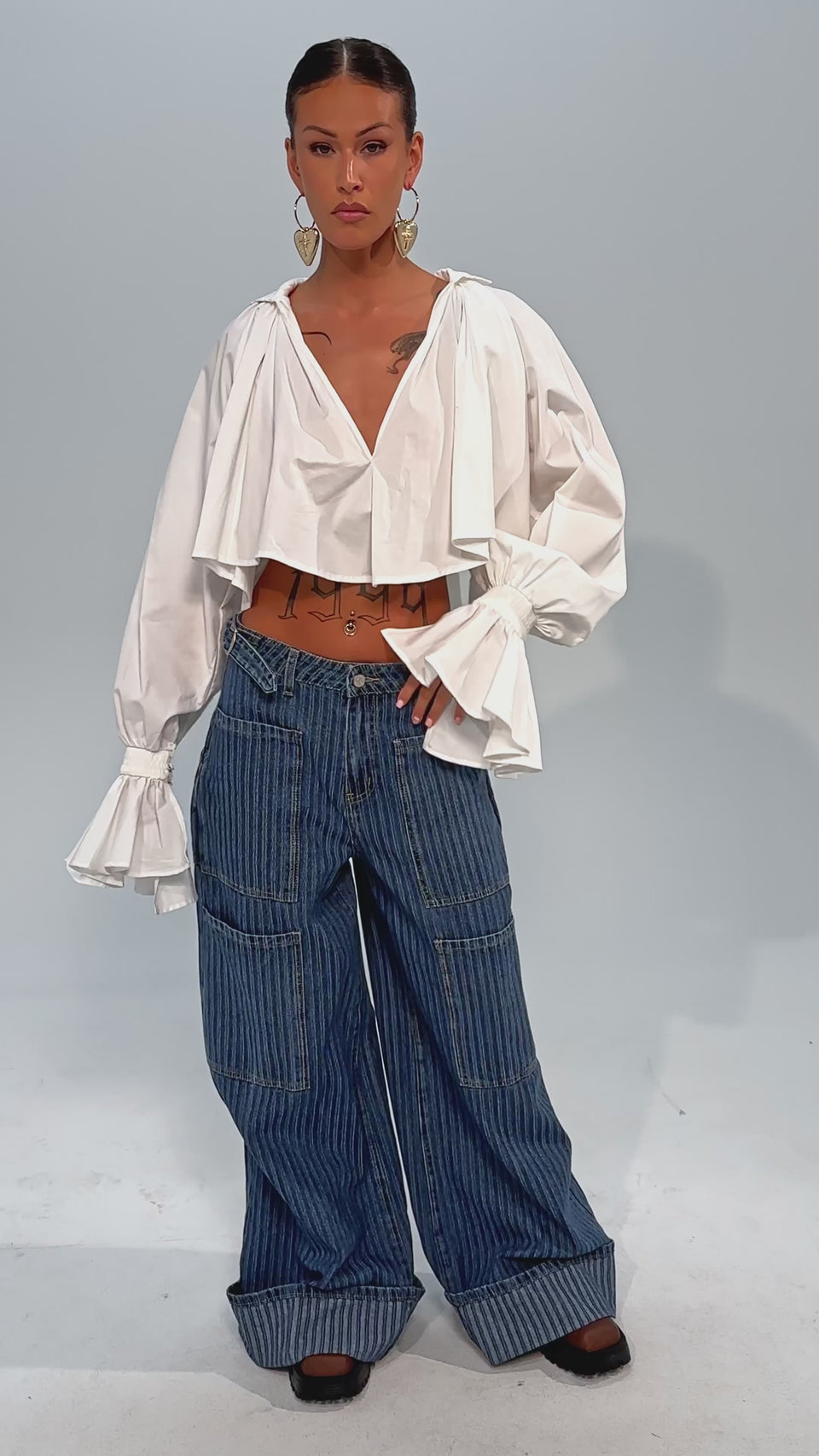 Video of woman modeling the BE MINE STRIPE DENIM wide-leg, high-rise jeans paired with a loose white blouse, showcasing a "1999" tattoo on her stomach. She accessorizes with hoop earrings and stands against a plain light background, hair tied back.
