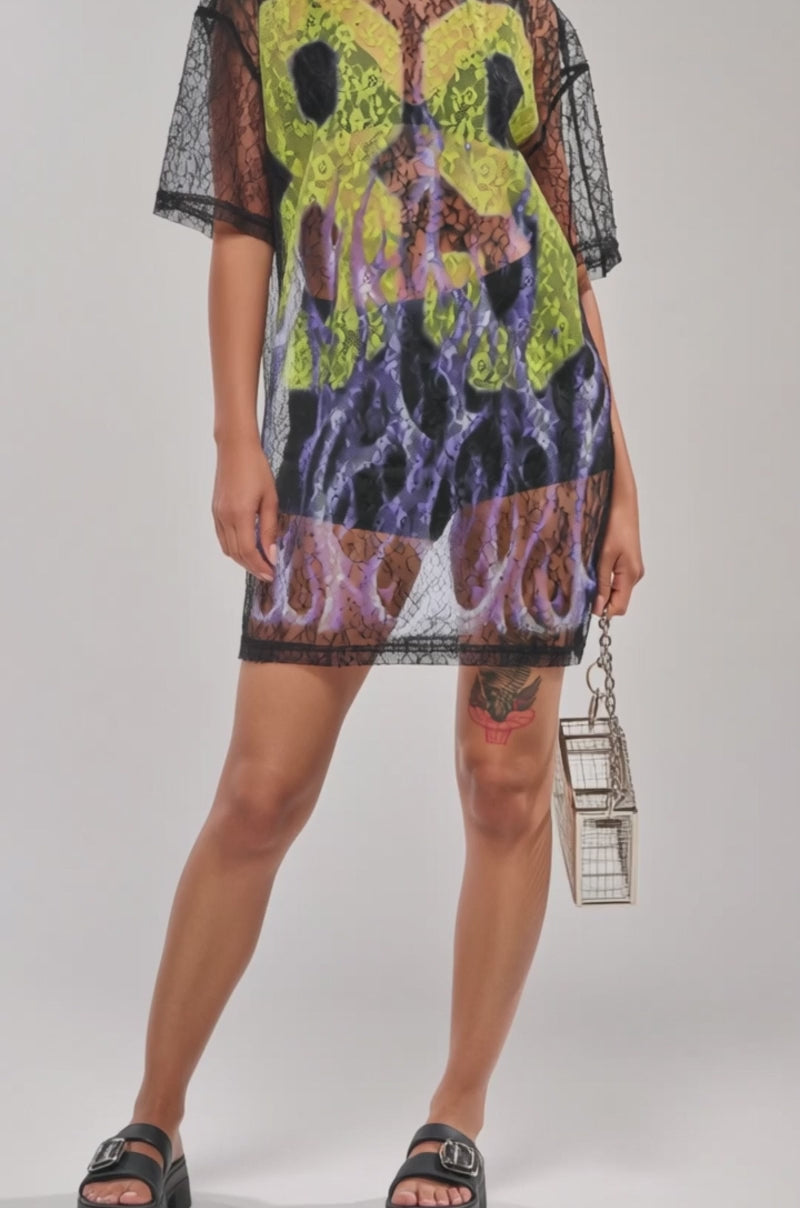 A video of a woman wearing sunglasses, a person models the ALL THE RAGE LACE JERSEY MINI DRESS—sheer black mesh with yellow and purple abstract patterns.