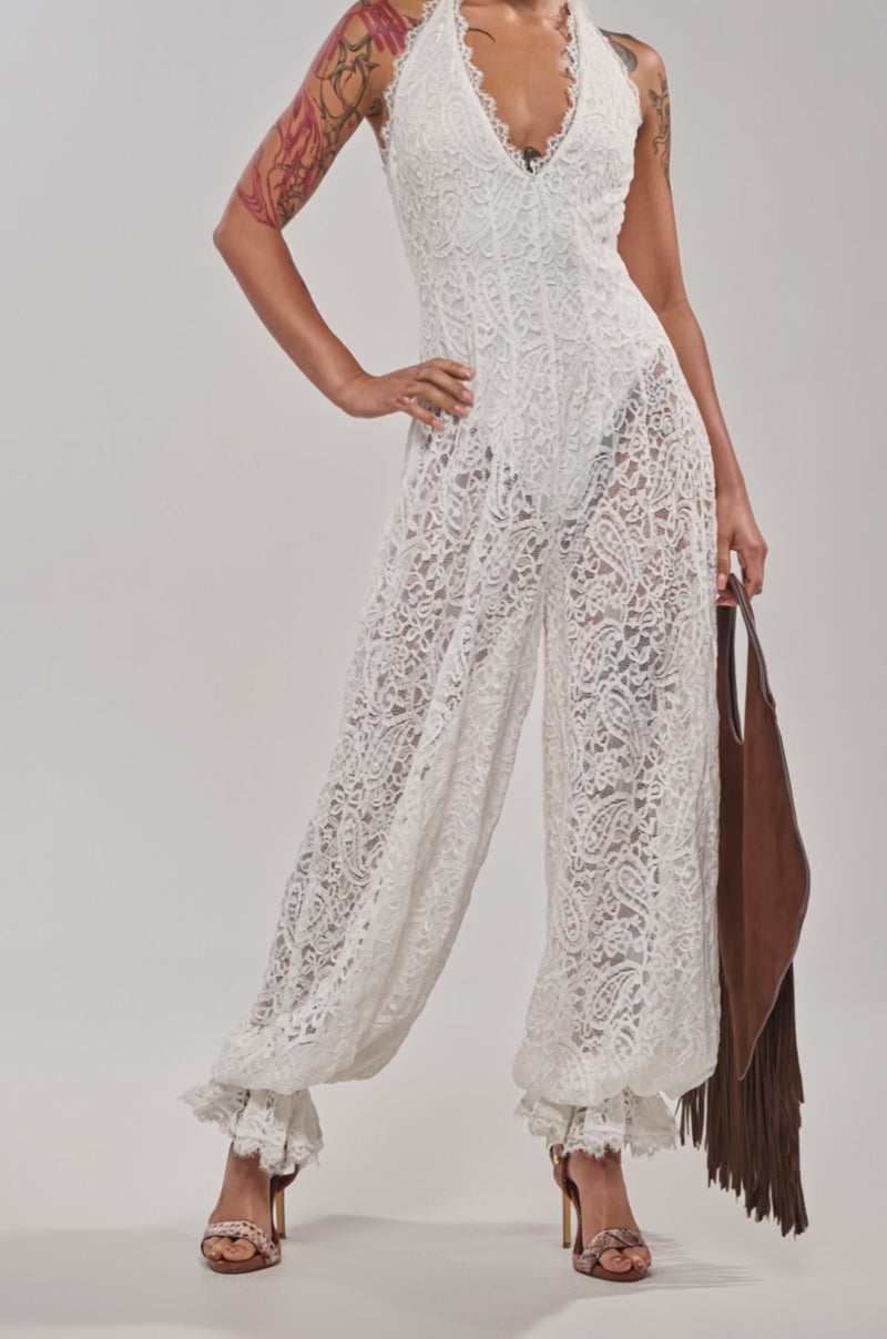 Video of woman modeling the GALAVANTING LACE JUMPSUIT IN IVORY—sleeveless, with ruffle ankle cuffs and a white belt—paired with hoop earrings and high heels. Her tattoos stand out against the plain light background.
