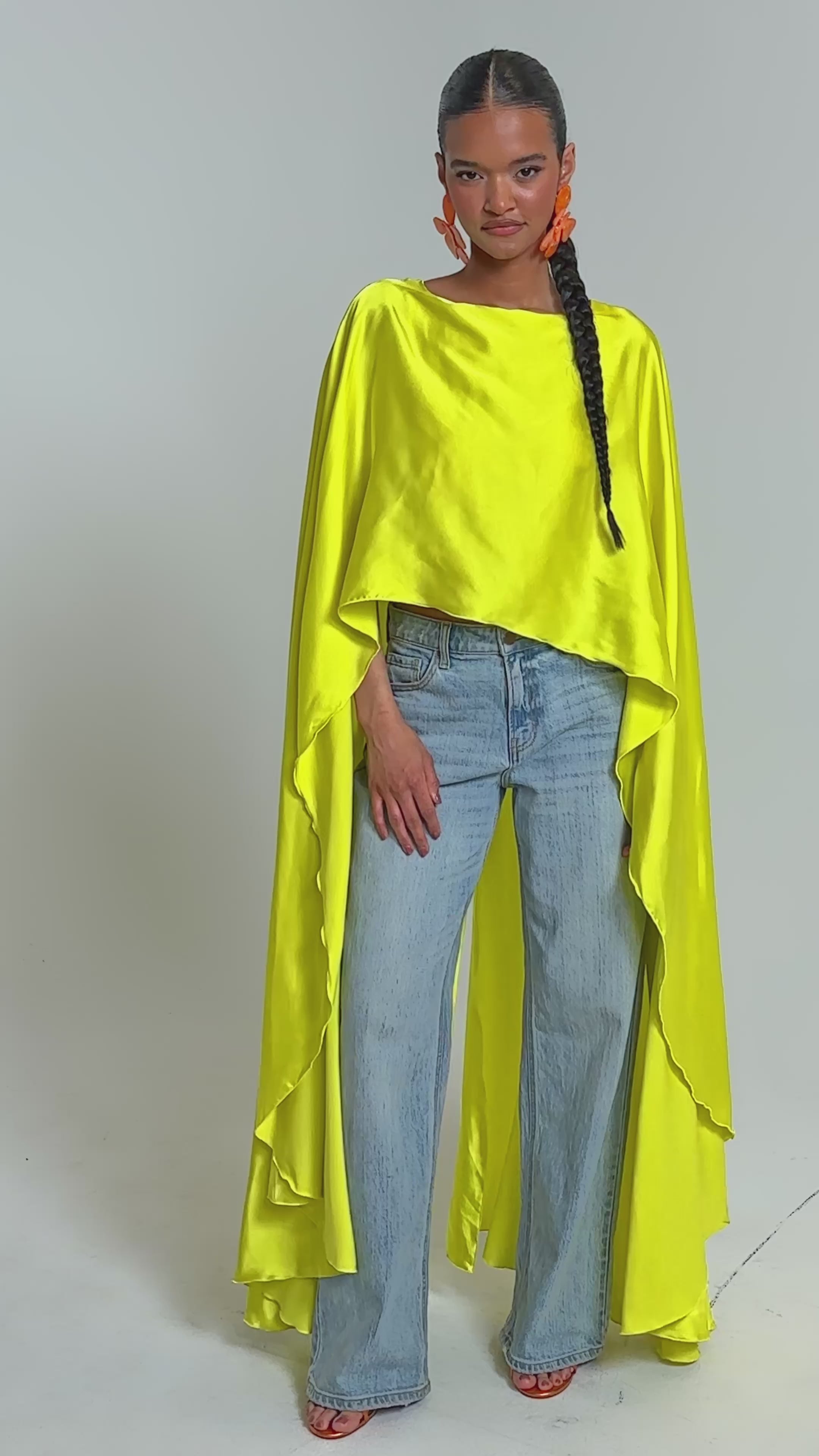 Video of woman modeling the SO CHIC CAPE BLOUSE IN YELLOW, light blue wide-leg jeans, orange flower-shaped earrings, and sandals stands confidently against a plain light background.