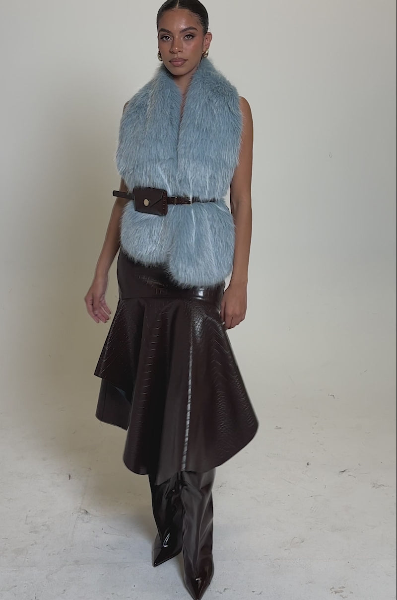 A video of a woman wearing the ITS GIVING FUR BLOUSE, a sleeveless light blue faux fur vest with an adjustable waist belt, paired with a dark brown textured asymmetrical midi skirt and brown knee-high boots.