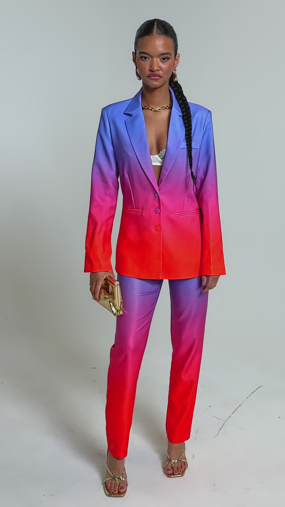 Video of woman modeling the SUNSET IN PARADISE OMBRE BLAZER with matching pants, a white lace bra, gold hoop earrings, a gold bracelet, and a shiny gold clutch against a plain background.