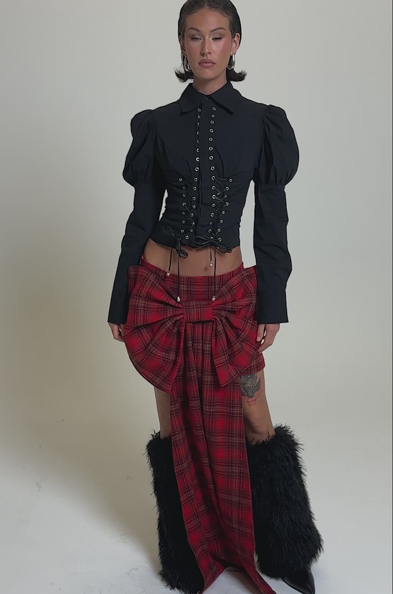 A video of a model posing against a plain background in the LOVELY DAYS PLAID BOW MINI SKIRT, styled with a fitted black button-up top with puffed sleeves and black furry boots.