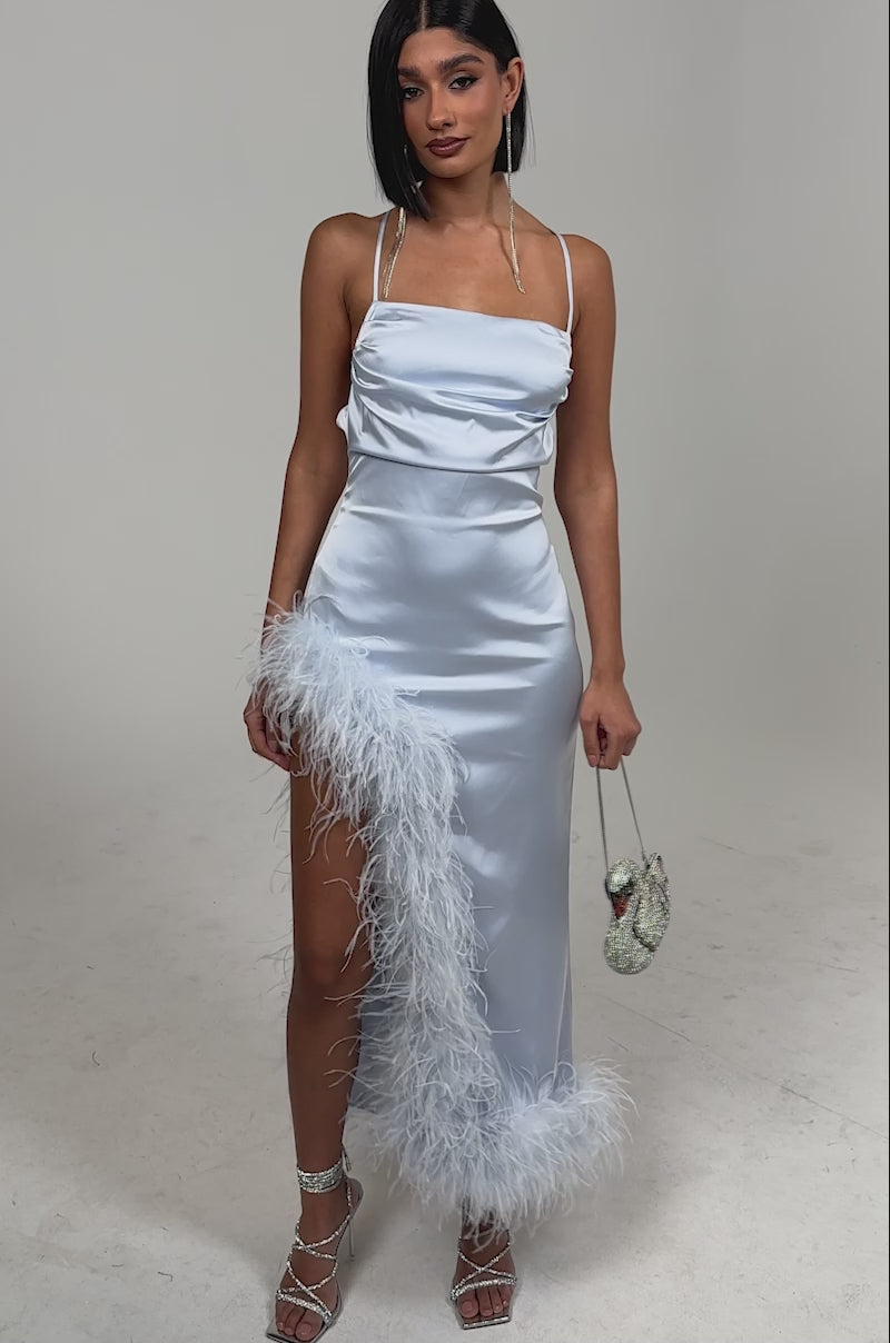 Video of woman modeling the FROSTBITE MAXI DRESS, a sleek gala dress with thin straps and feather-trimmed high slit, paired with strappy heels and a silver purse against a plain light background.