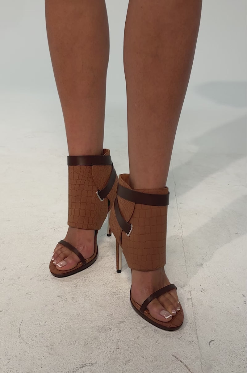 A video of a model wearing the Azalea Wang ELMER-BROWN sandal. Crafted from croc embossed brown faux leather, these striking sandals feature an open almond toe with a thin toe strap, a slim stiletto heel, a Comfort Cushion insole, and a split ankle-high shaft with an adjustable wraparound belt for a customizable fit.