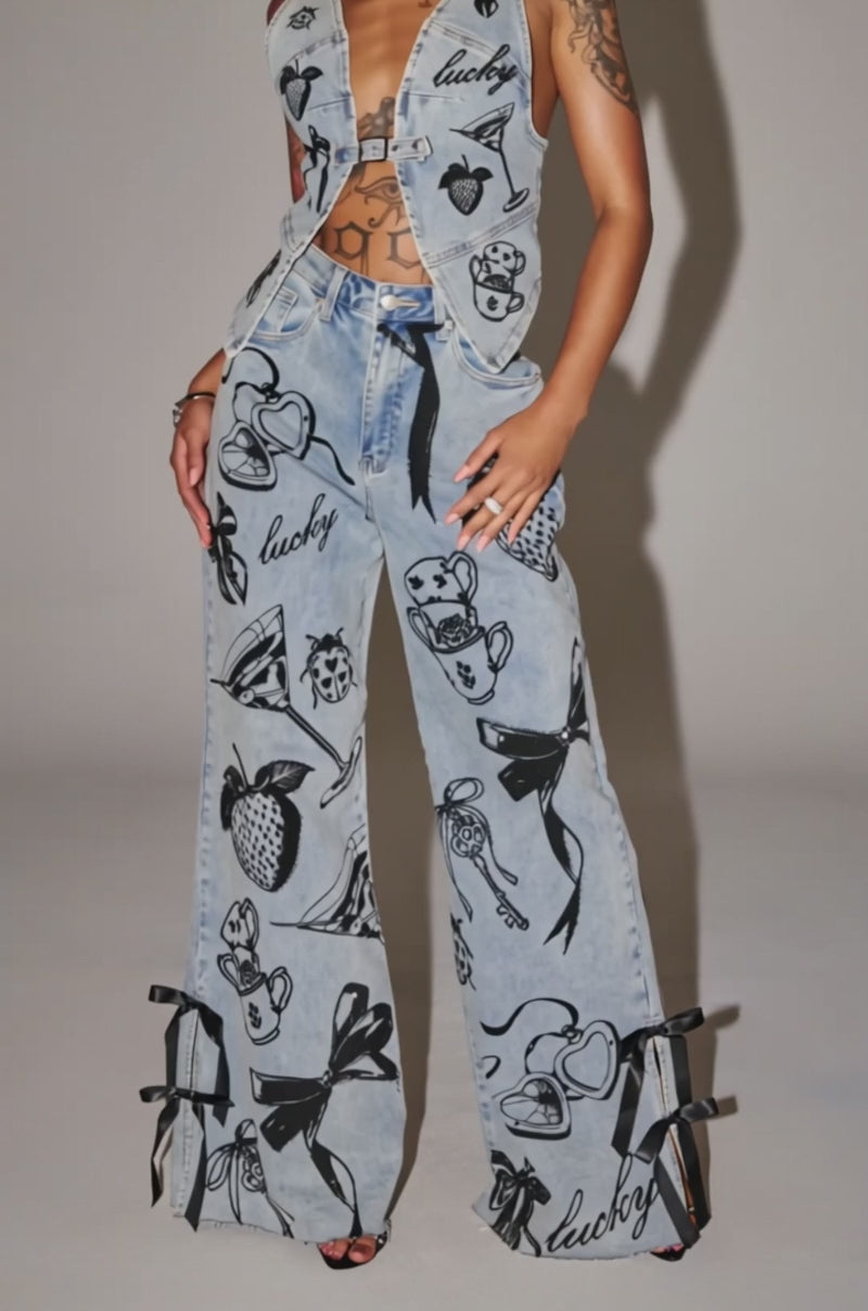 Video of woman modeling the AFTER HOURS STRAIGHT LEG DENIM, light blue jeans featuring black sketch graphics of hearts, strawberries, ladybugs, teacups, ribbons, and "lucky," with black ribbon bows accenting each leg's hem.