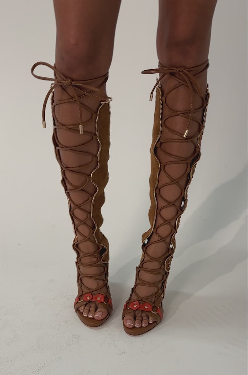 Video of woman modeling the AZALEA WANG SHAI TAN GLADIATOR STILETTO SANDAL—knee-high heels with intricate red and brown cut-out designs and a tan stiletto silhouette, set against a plain, light background.