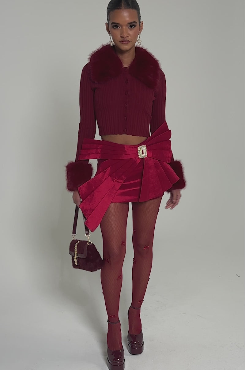 Video of woman modeling the IMPRESSIVE SKIRT featuring pleats and a jeweled belt, paired with a cropped sweater with fur cuffs, sheer bow-detailed tights, platform heels, and a fuzzy red handbag.