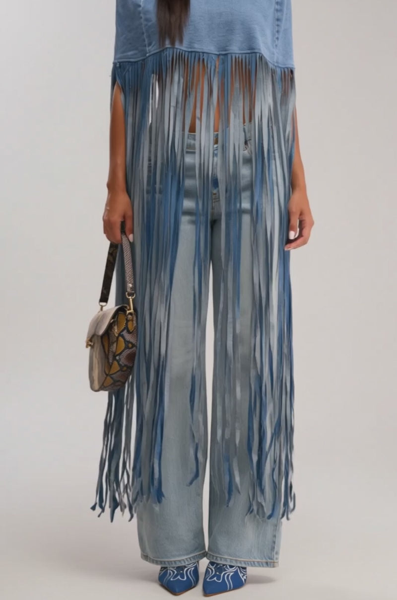 Video of woman modeling the DENIM DREAMS BLOUSE with wide-leg jeans and embroidered blue shoes, holding a small snakeskin-patterned handbag. Her long dark hair and the beige background highlight her chic western style.