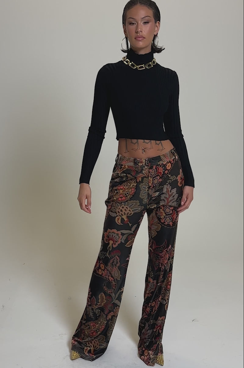 A video of a person in a black ribbed crop top and the FALLING IN LOVE FLORAL TROUSER—high-rise, flared pants with bold red, orange, and brown florals—finished with a chunky gold chain necklace.