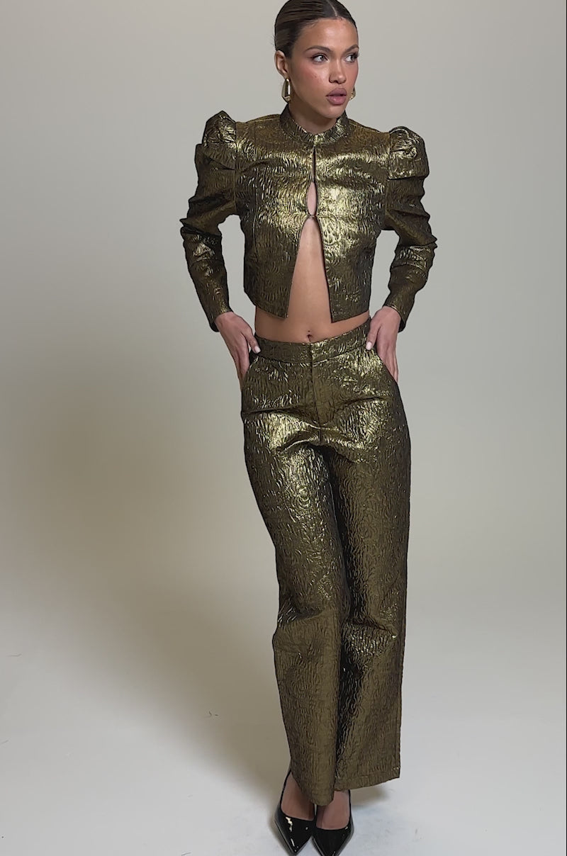 Video of woman modeling the DAPHNE BROCADE TROUSER and a matching jacket, with one hand in their pocket and pointed black high heels, a person stands against a plain light background.