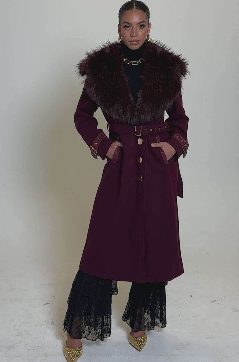 A video of a woman wearing the CATALINA FUR COLLAR TRENCH—a longline jacket with a large fur collar, gold buttons, and belt—styled with gold earrings, a black turtleneck, layered black lace skirt, and gold heels.