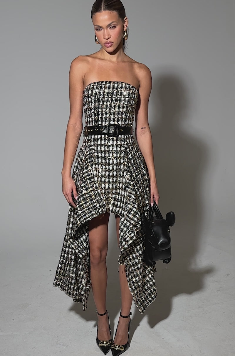 Video of woman modeling the PURE ROYALTY BROCADE HIGH LOW TWEED DRESS WITH SEQUINS—strapless, high-low, and cinched with a black belt—styled with gold heels, hoop earrings, and a black handbag against a plain gray backdrop.