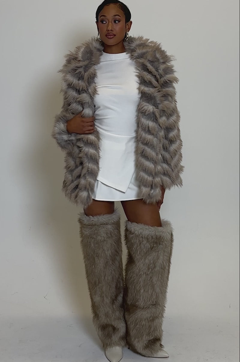 Video of woman modeling the SHOWBIZ BABY FAUX FUR, a fluffy chevron print coat with an oversized collar, paired with a white mini skirt and matching fur leg warmers, posing against a plain light background.