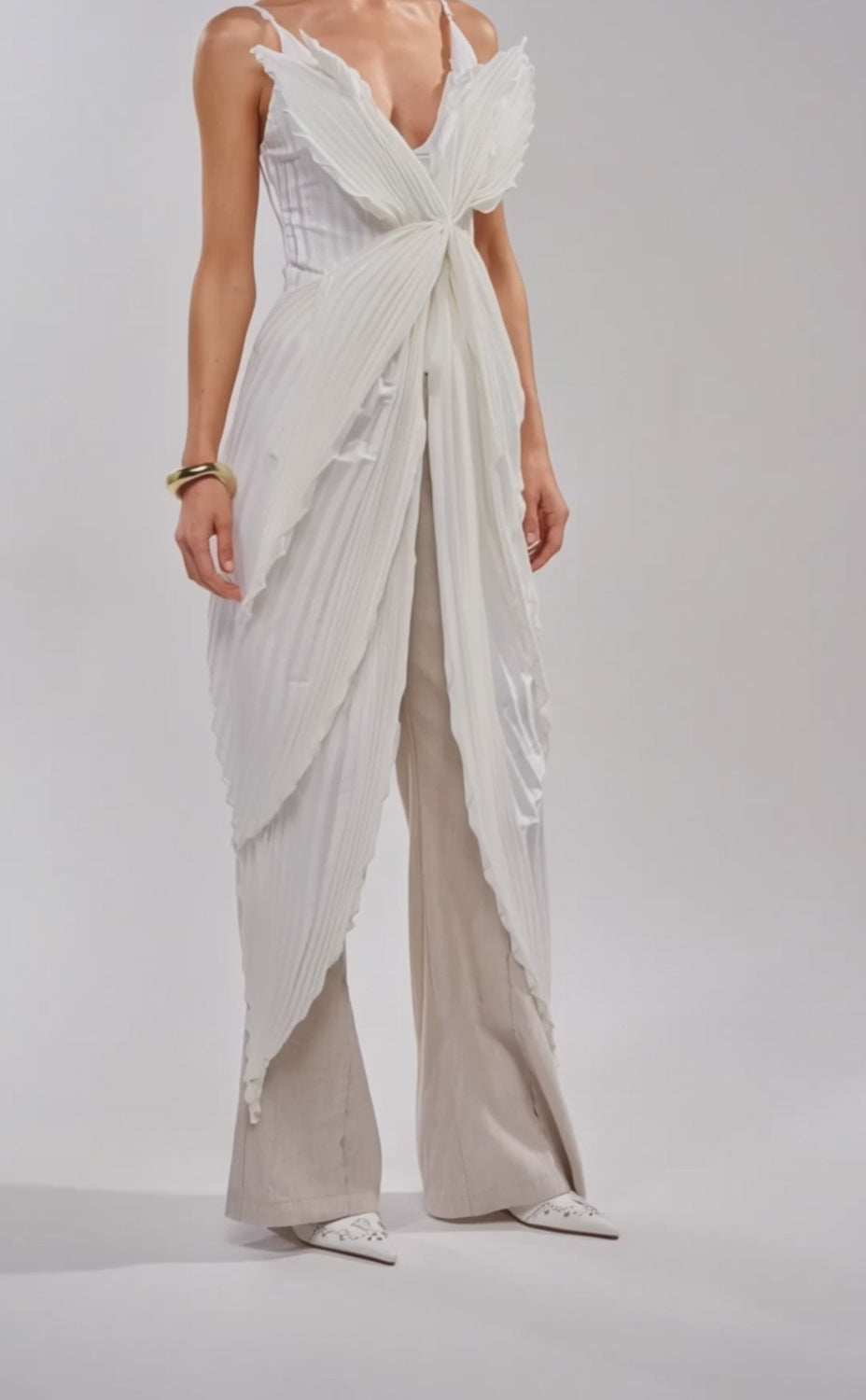 A video of a woman in the FLYING FREE LAYERED TOP—satin white with pleated panels and a large front bow—paired with loose beige pants and white pointed-toe heels.