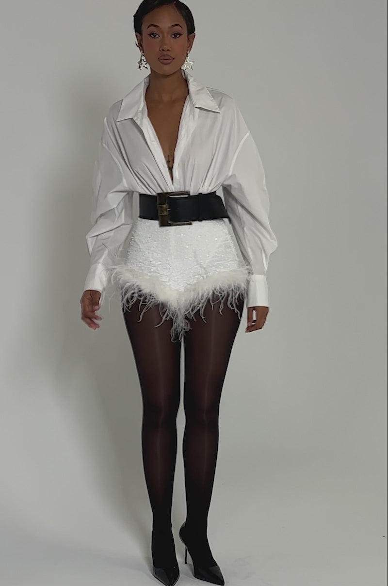 Video of woman modeling the NEW YEAR FEATHER SHORTS—high-rise mini shorts embellished with sequins and feather trim at the hem—paired with a tucked-in white, long-sleeved blouse.