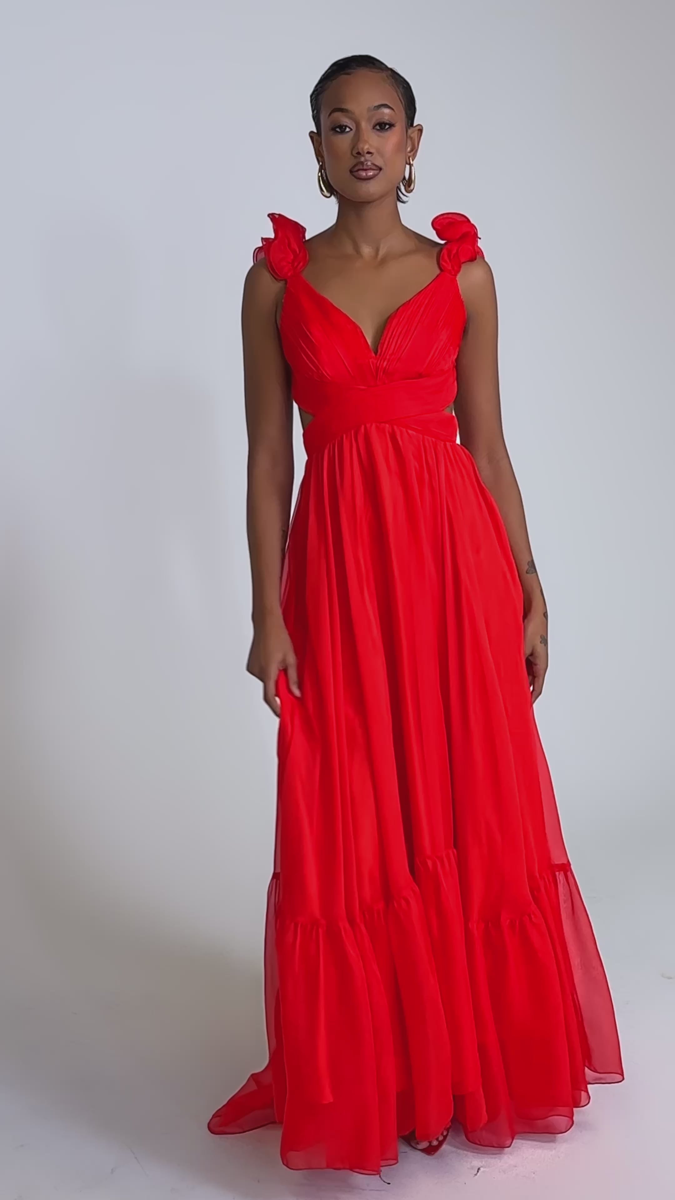 A video of a woman in the DREAM GIRL MAXI DRESS IN RED, featuring ruffle shoulder details and light, airy fabric, standing confidently against a white background, highlighting the dress’s elegant, flowing full-length silhouette.