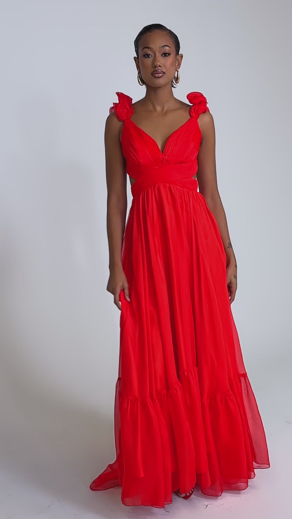 A video of a woman in the DREAM GIRL MAXI DRESS IN RED, featuring ruffle shoulder details and light, airy fabric, standing confidently against a white background, highlighting the dress’s elegant, flowing full-length silhouette.