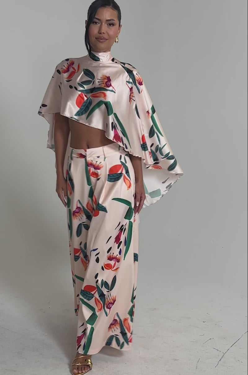Video of woman modeling the CANDY RAIN SATIN WIDE LEG PANT, featuring vibrant multicolored floral and leaf prints. She pairs it with a light pink crop top, gold sandals, gold earrings, and wears her hair partially tied back.