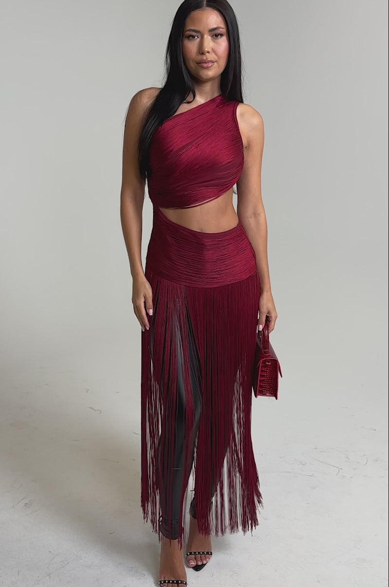 A video of a woman wearing the LOVE STRUCK FRINGE TOP in burgundy, styled with black leather leggings, strappy heels, and a small matching burgundy handbag.