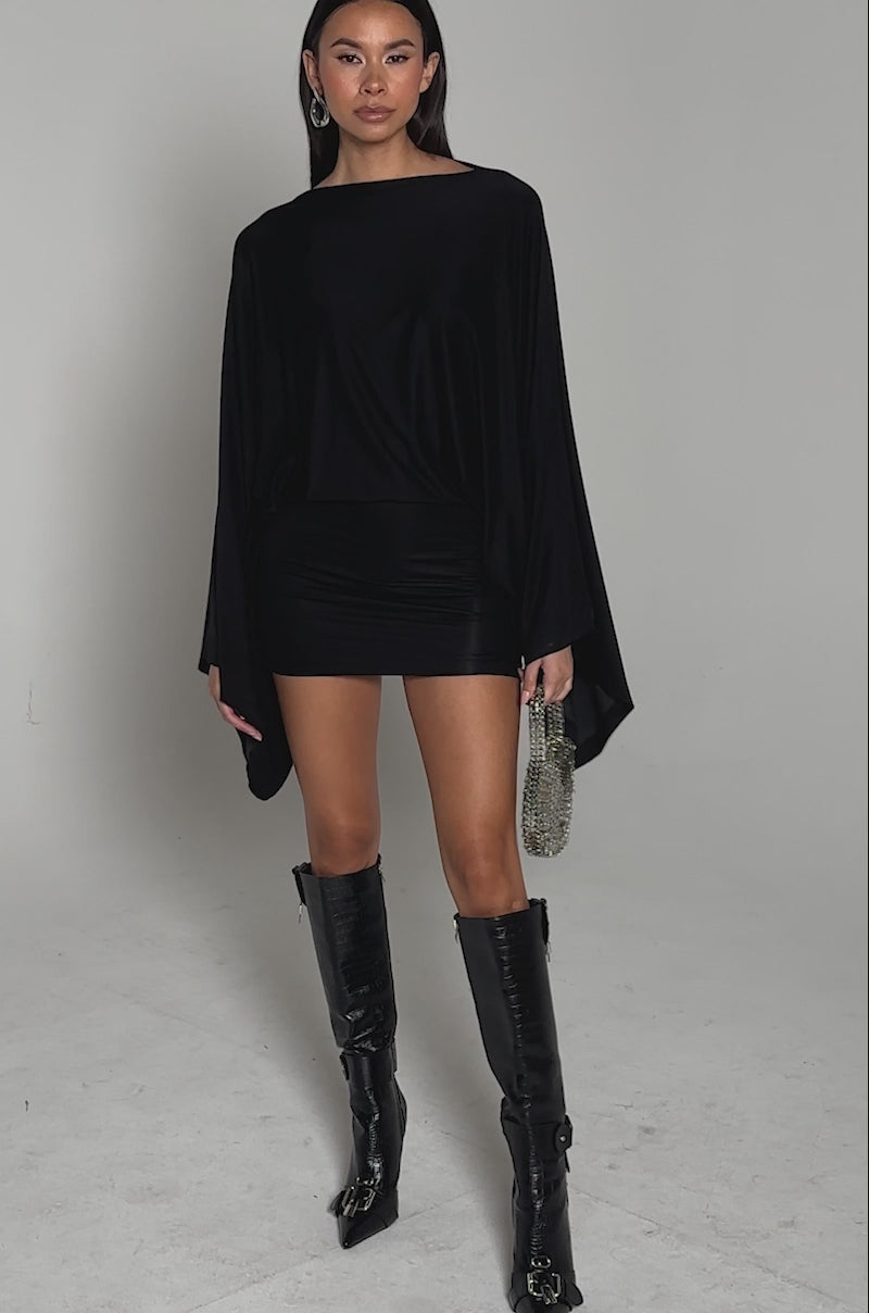 Video of woman modeling the DIANA DRAPED MINI DRESS IN BLACK, featuring batwing sleeves and a fitted mini skirt, paired with a shiny silver purse, posed against a plain light background.