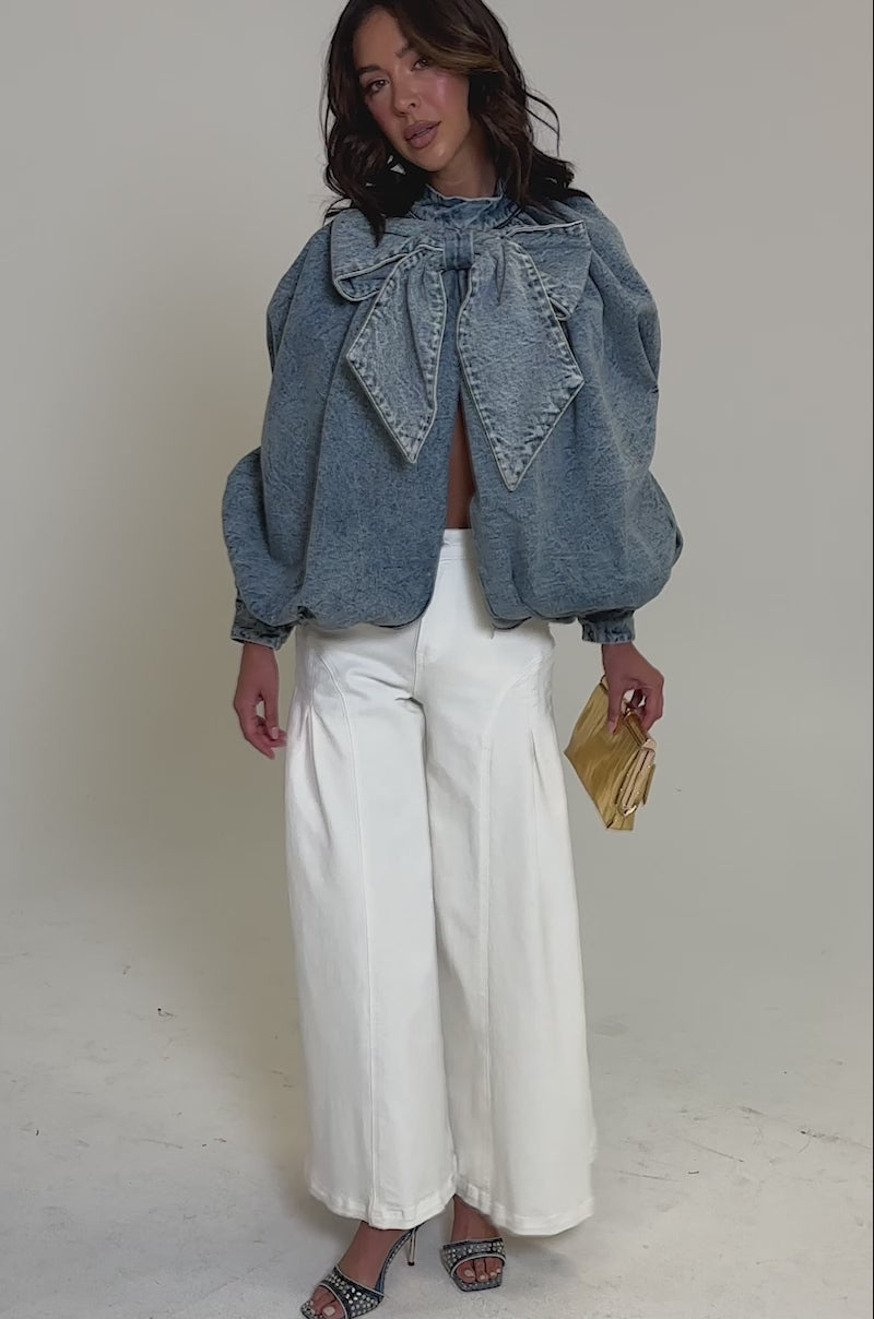 Video of woman modeling the NEW DREAM PUFF SLEEVE TOP, a light-wash denim piece with an oversized collar bow and billowy sleeves, paired with high-waisted white pants. She carries a small gold handbag and has loose, wavy hair.