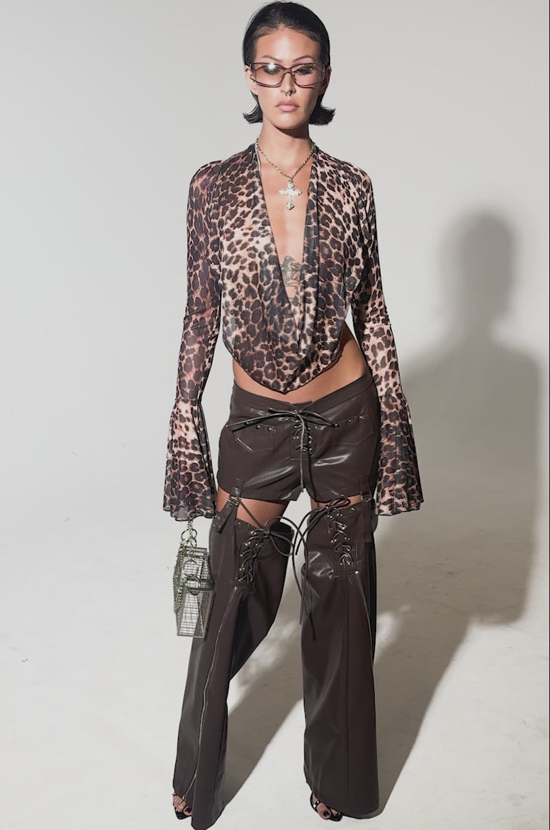 A video of a person wearing festival-ready style in WANNA BE FAUX LEATHER CHAPS, paired with a sheer leopard-print blouse, lace-up brown mini skirt, black heels, oversized sunglasses, and a silver handbag for a confident look.