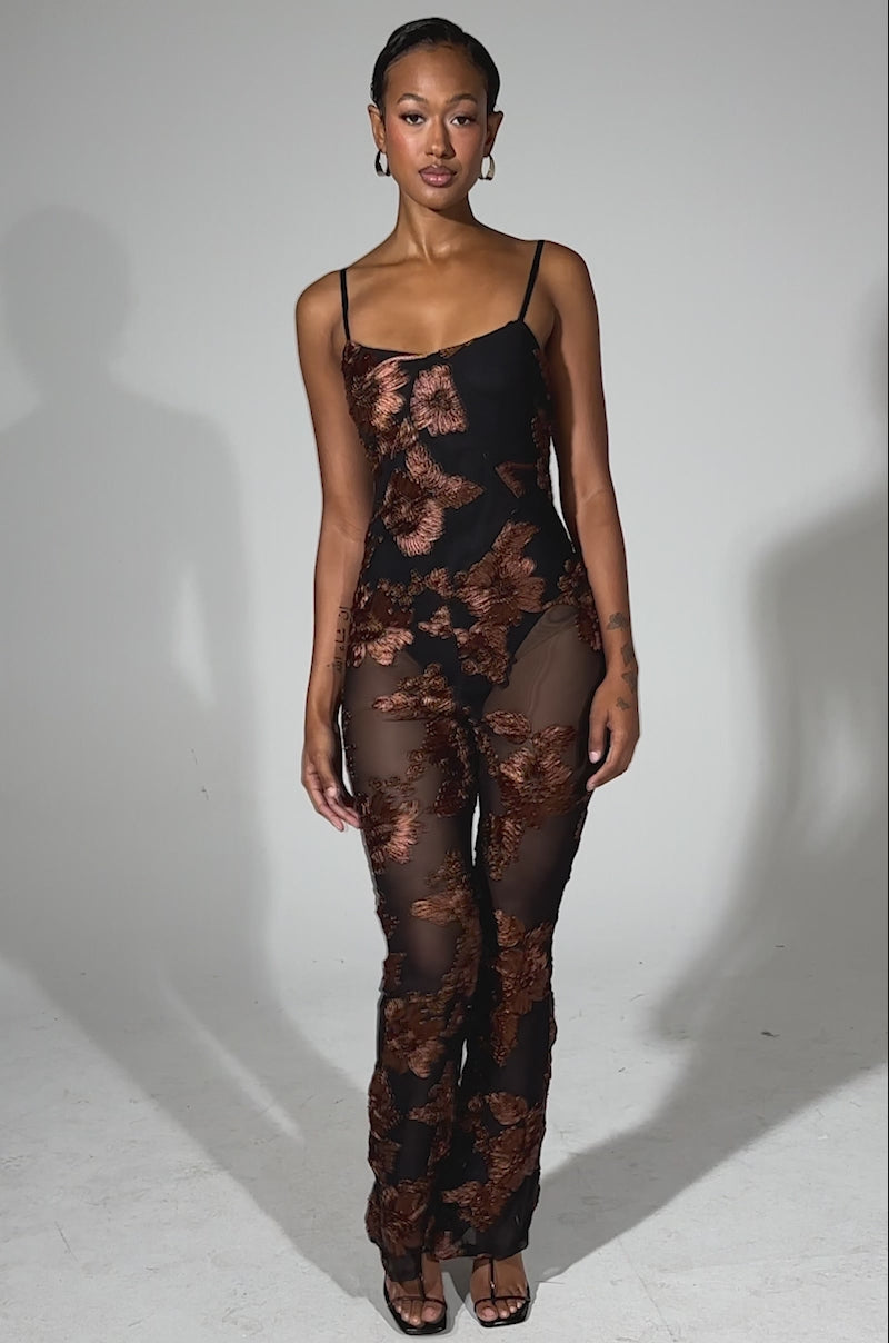 A video of a woman posing facing forward in the LAY IT LOW PRINTED MESH JUMPSUIT, a sheer black jumpsuit with bold brown floral prints, paired with black heels and hoop earrings against a plain background.