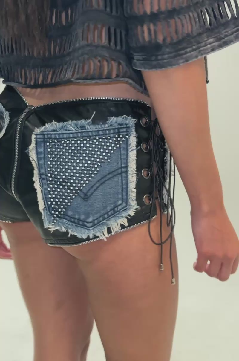 Video of a woman modeling the HOT AND DANGEROUS HOT SHORT, a person shows off high-waisted shorts crafted with bold patchwork construction that merges black and blue denim panels, leather accents, zippers, studs, grommets, and frayed edges.