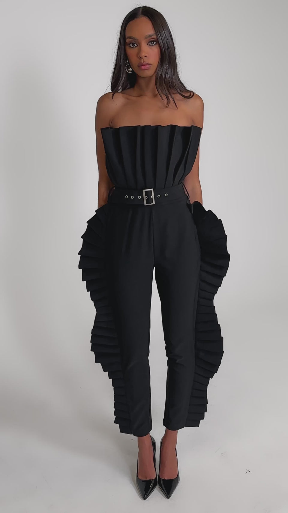 Video of woman modeling the RUFFLE MY FEATHERS BLACK JUMPSUIT, featuring dramatic pleated side ruffles and a strapless design, with pointed black heels 