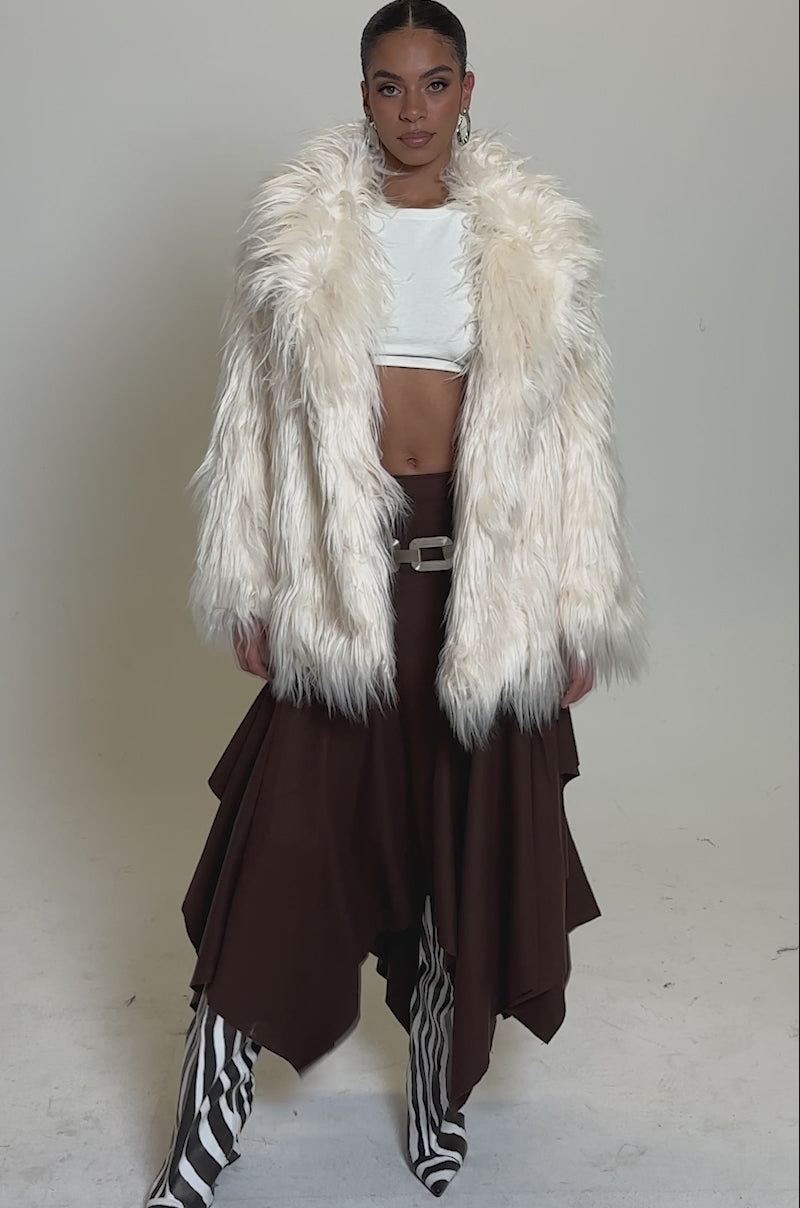A video of a woman in the PARK CITY FAUX FUR COAT over a white top, layered brown skirt, and bold black-and-white zebra-striped boots against a plain light background.