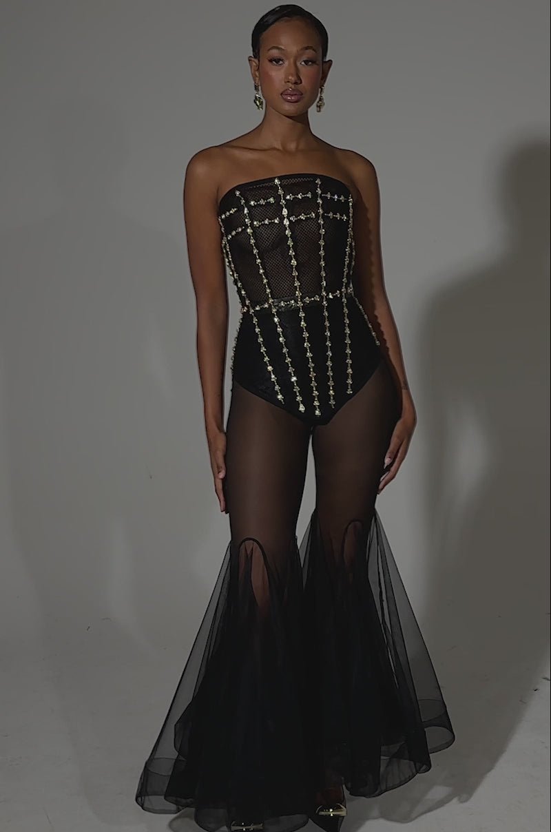 A video of a woman wearing the SMOKESHOW CORSET WITH RHINESTONES, a strapless black bodysuit with gold embellishments, paired with sheer black flared tulle pants, black pointed heels, and gold earrings against a plain light background.