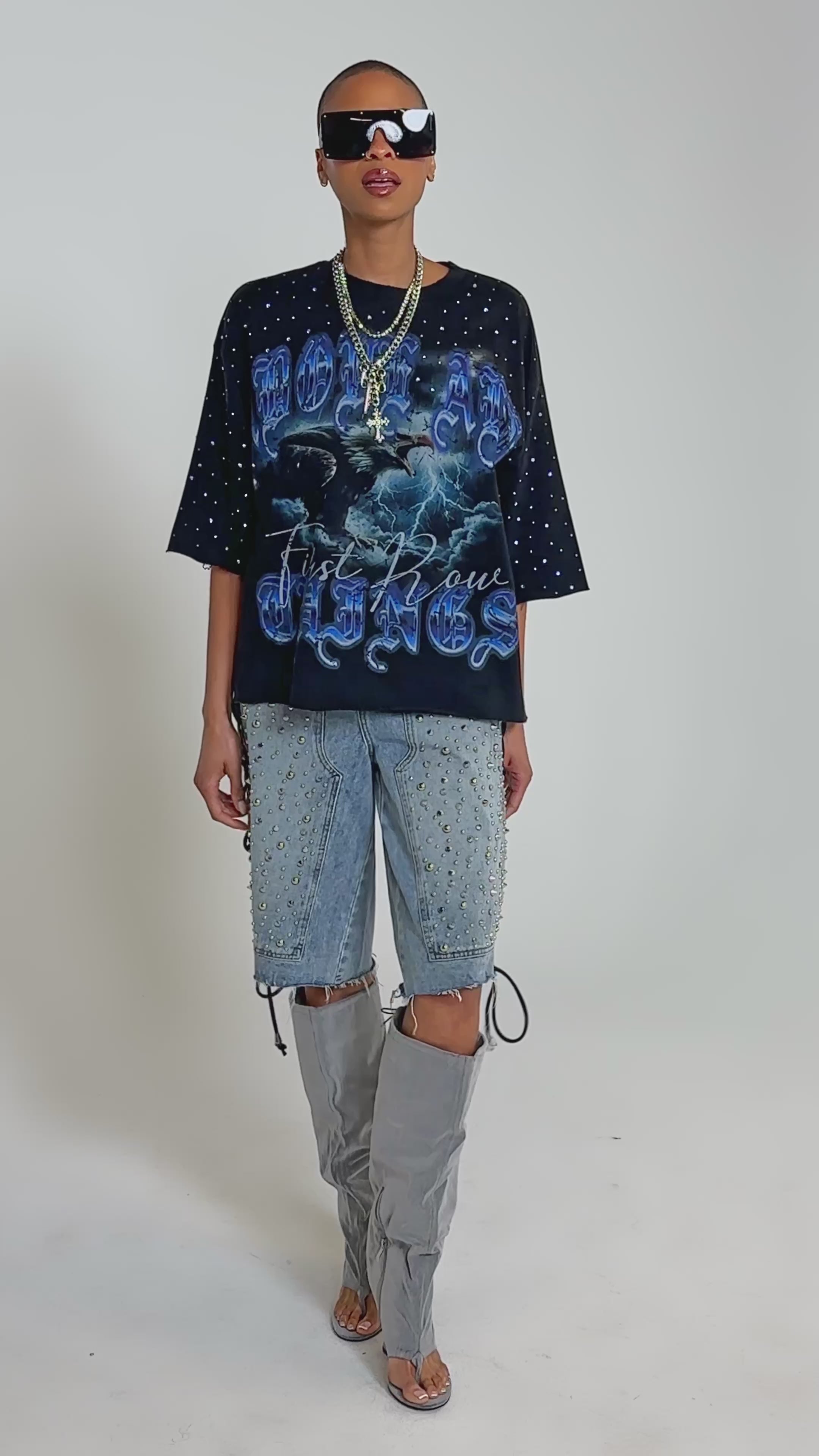 Video of woman modeling the FIRST ROW TAKING FLIGHT FRENCH TERRY TEE with blue gothic lettering, layered silver necklaces, and studded denim shorts, standing against a plain background.