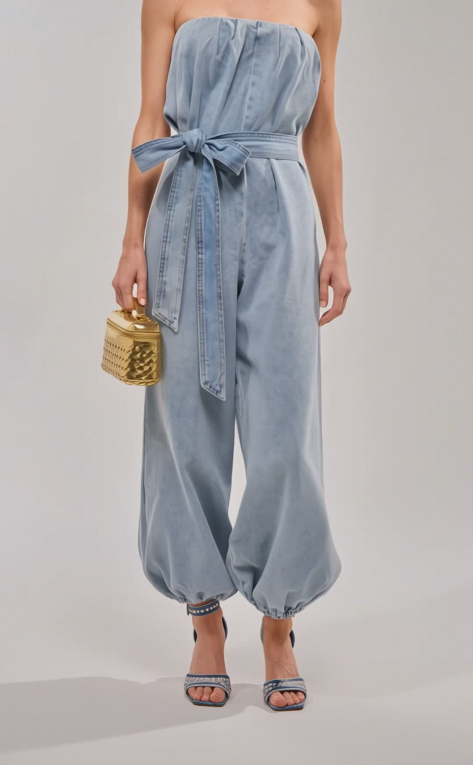 Video of woman modeling the BREEZY DAY DENIM JUMPSUIT in light-wash denim with a strapless top, wide fabric belt, and balloon pant legs. She pairs it with a gold handbag and blue strappy heels against a plain light gray background.