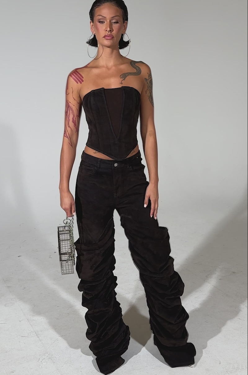A video of a woman posing confidently in the OH BABY VELVET DENIM PANT, a strapless, dark brown jumpsuit with high-rise relaxed fit and ruched wide-leg pants. She holds a small silver handbag, her tattoos showing and hair slicked back.