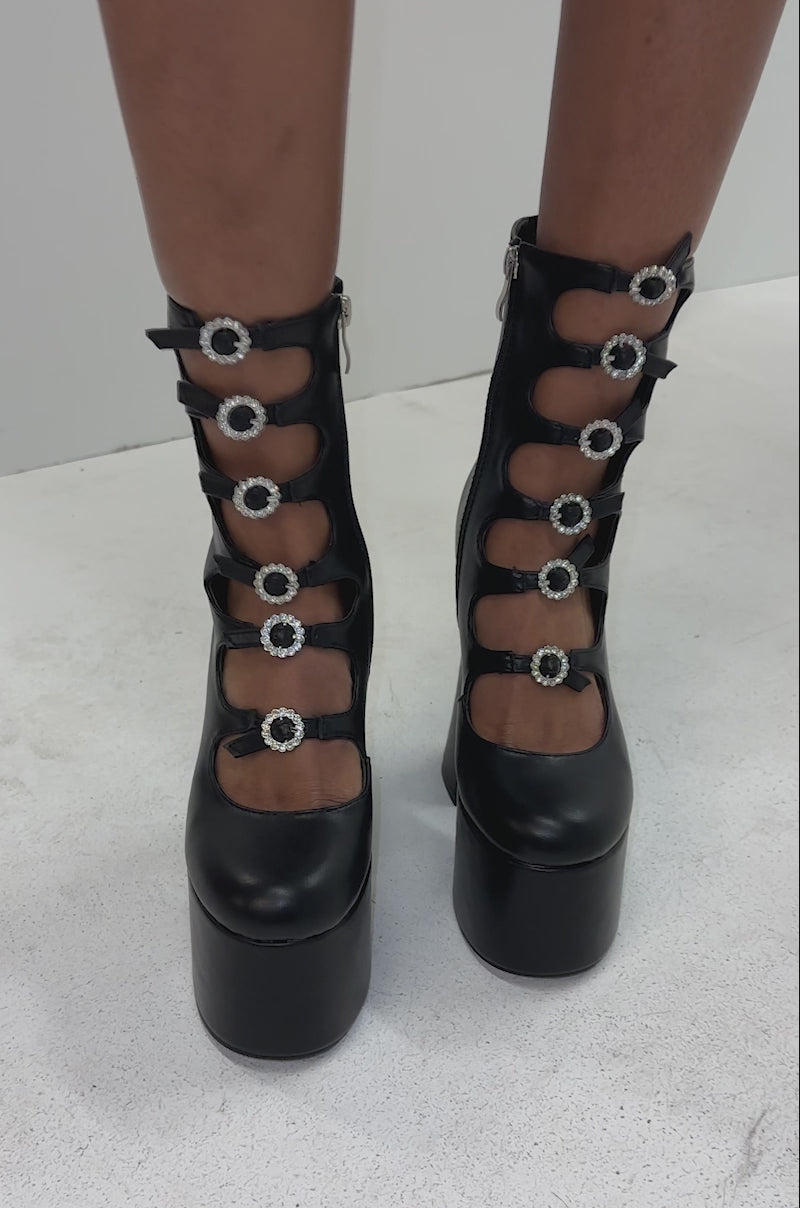 A video of a person wearing the AZALEA WANG OMLEE BLACK ULTRA HIGH PLATFORM BOOTIE, featuring chunky heels and multiple buckle straps, standing against a white background.