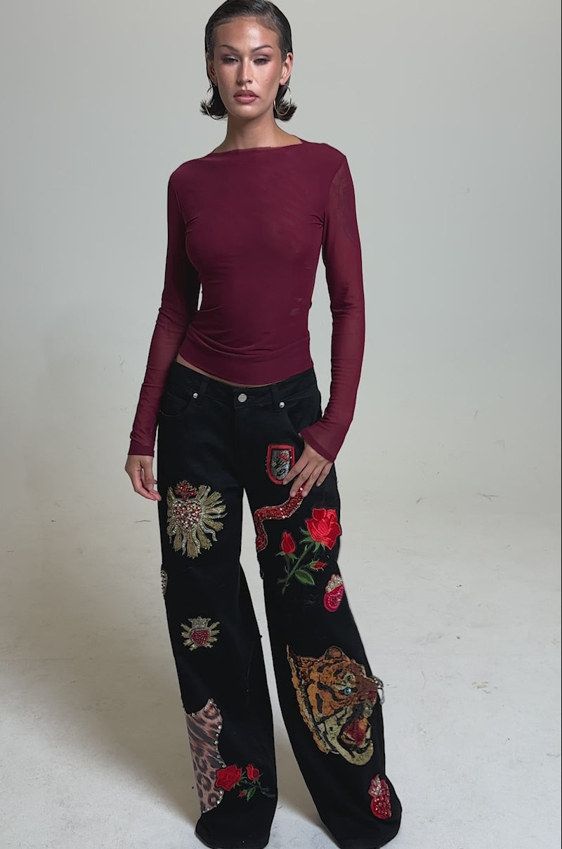 A video of a woman in the PAXTON Double Lined Mesh Long Sleeve Top in Plum, paired with high-waisted black jeans with bold animal and floral embroidery. 