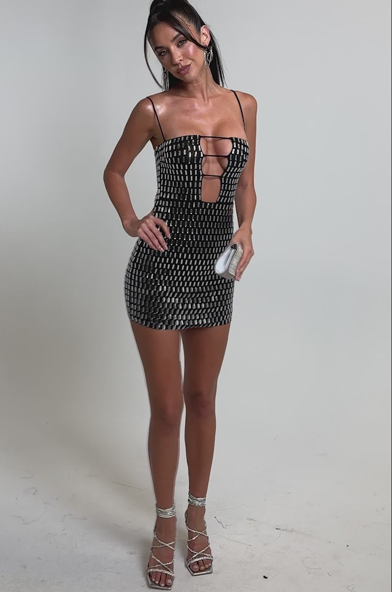 A video of a woman wearing the HARD NOT TO STARE RHINESTONE MINI DRESS, styled with silver high-heeled sandals, hoop earrings, and a silver clutch.